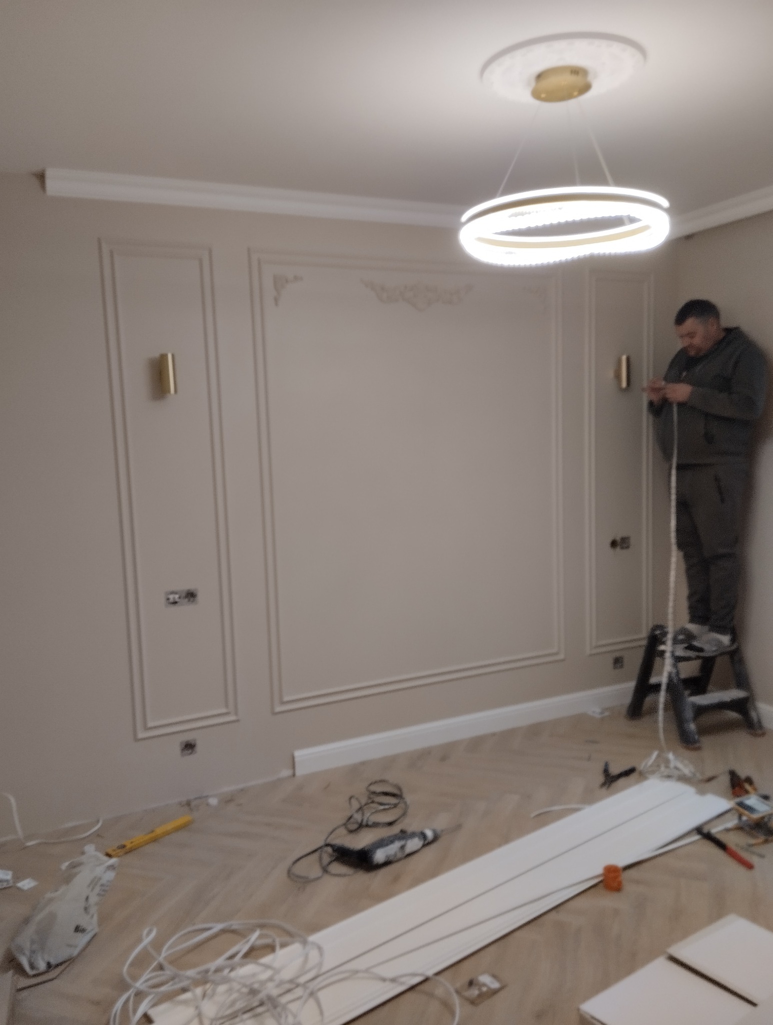 Instalacja listew dekoracyjnych na ścianie z ozdobnymi panelami; pracownik na drabinie, widoczne narzędzia i materiały na parkiecie w jodełkę, nowoczesna lampa sufitowa.