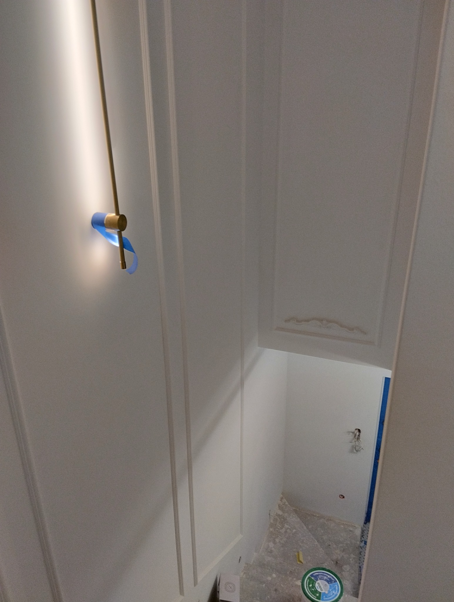 Świeżo pomalowana, biała klatka schodowa z dekoracyjnymi listwami przyściennymi, zabezpieczona niebieską taśmą malarską wokół nowoczesnej, złotej lampy ściennej. Schody w trakcie przygotowania...