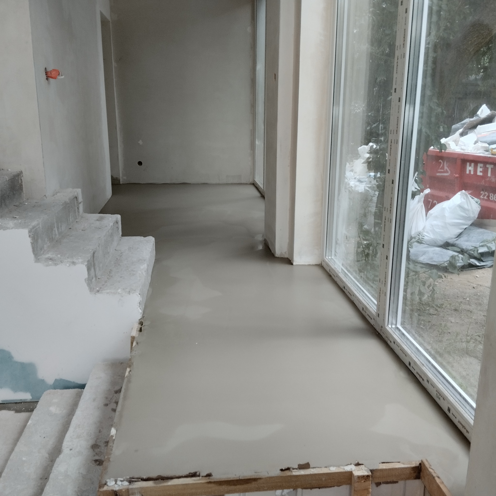 Wylewka samopoziomująca na podłodze w surowym wnętrzu, widoczne schody w stanie deweloperskim i okno z widokiem na zewnątrz, w tle kontener na gruz.
