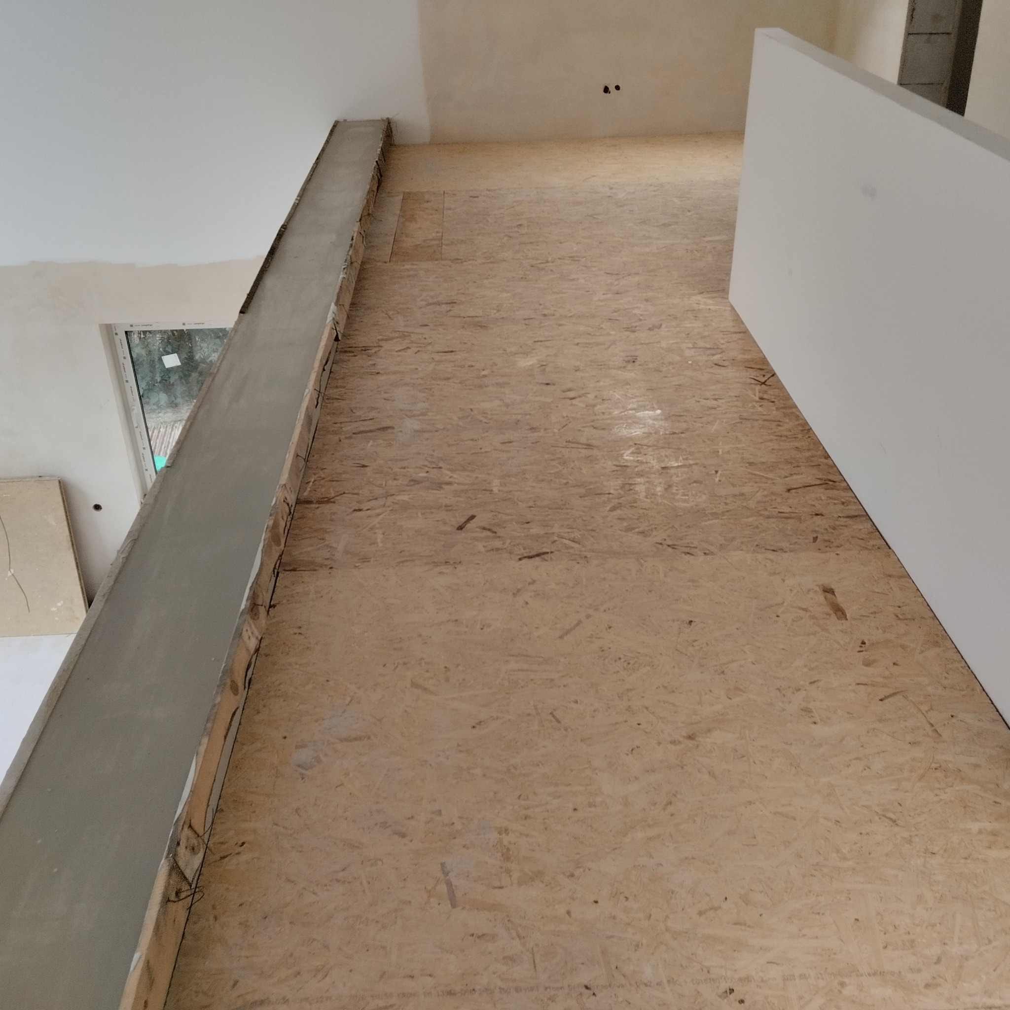 Surowe wnętrze w trakcie prac wykończeniowych: podłoga z płyt OSB, ściany przygotowane pod malowanie, widoczna płyta karton gips i element konstrukcyjny z betonu.