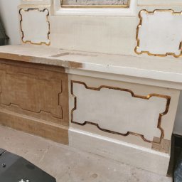 Czyszczenie Laserem - Fragment zabytkowego mebla sakralnego w trakcie renowacji: połowa powierzchni oczyszczona z farby, ukazująca naturalne drewno, druga połowa z białym wykończeniem i złotymi zdobieniami.