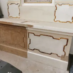 Fragment zabytkowego mebla sakralnego w trakcie renowacji: połowa powierzchni oczyszczona z farby, ukazująca naturalne drewno, druga połowa z białym wykończeniem i złotymi zdobieniami.