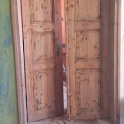 Drewniane, dwuskrzydłowe drzwi po renowacji, z widoczną strukturą drewna i śladami wcześniejszych warstw farby, częściowo otwarte, z kablem elektrycznym i taśmą ostrzegawczą na podłodze w tle...