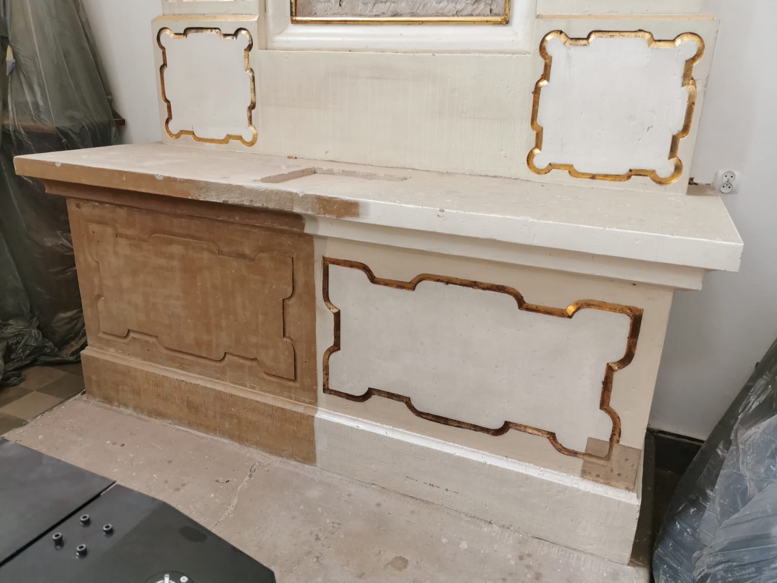 Fragment zabytkowego mebla sakralnego w trakcie renowacji: połowa powierzchni oczyszczona z farby, ukazująca naturalne drewno, druga połowa z białym wykończeniem i złotymi zdobieniami.