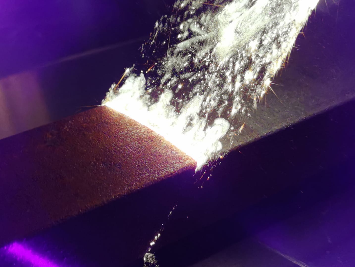 Strumień ścierniwa pod ciśnieniem uderza w rdzewiejący metalowy element, usuwając korozję i zanieczyszczenia. Widoczne iskry i pył.