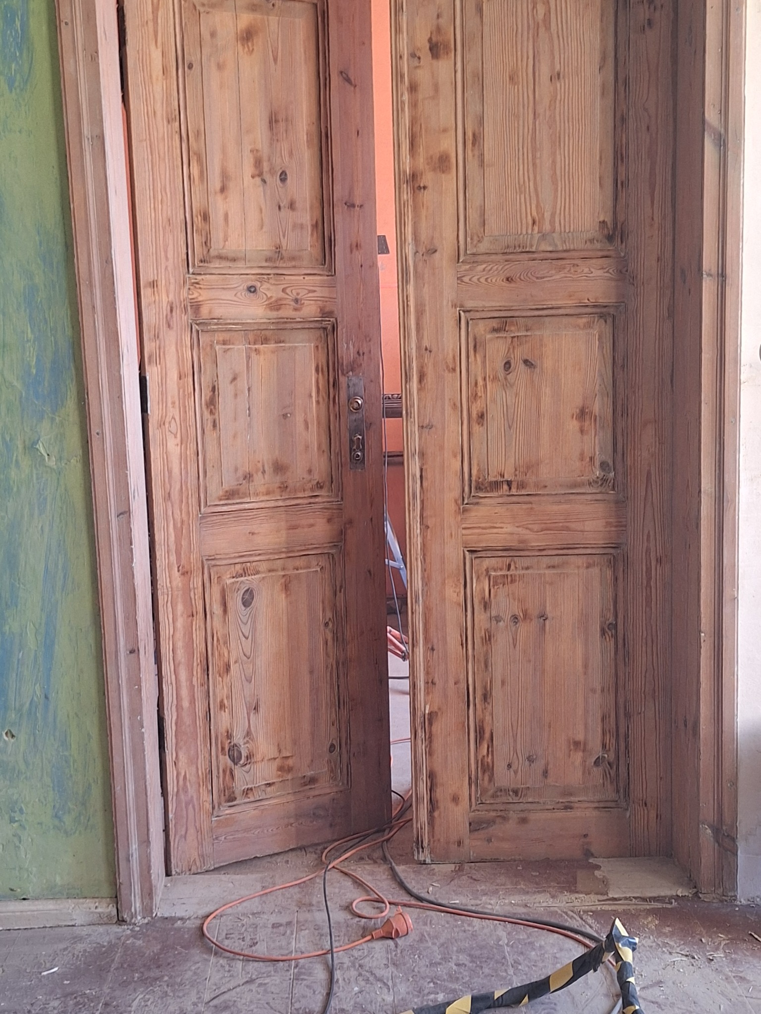 Drewniane, dwuskrzydłowe drzwi po renowacji, z widoczną strukturą drewna i śladami wcześniejszych warstw farby, częściowo otwarte, z kablem elektrycznym i taśmą ostrzegawczą na podłodze w tle...