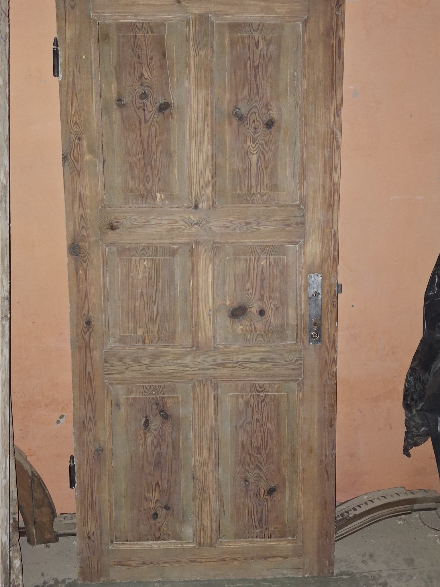 Stara, drewniana, sześciopanelowa drzwi po renowacji, z widocznymi śladami użytkowania i oryginalną, metalową klamką, oparta o pomarańczową ścianę w trakcie remontu.