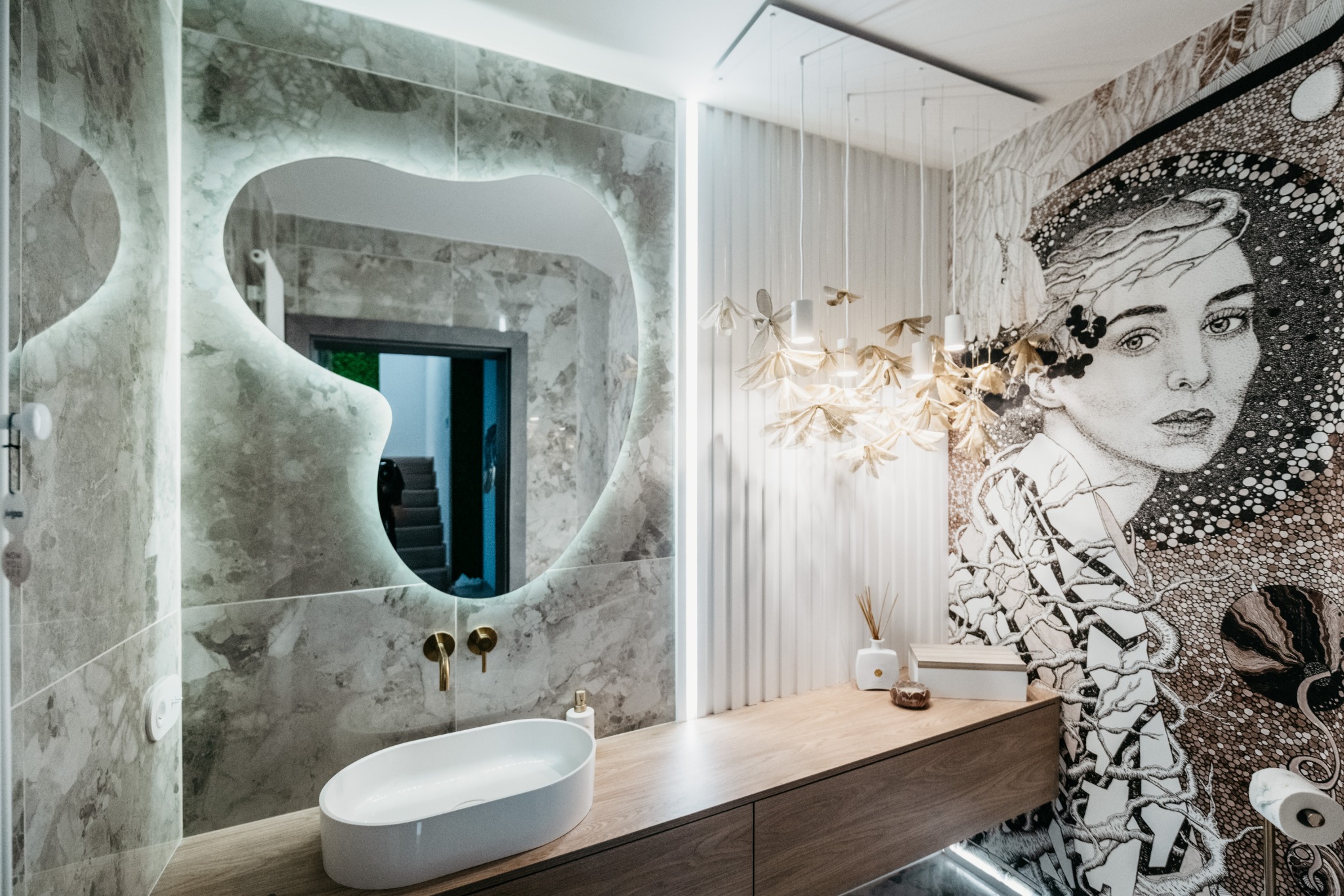 Nowoczesna łazienka z podświetlanym lustrem o nieregularnym kształcie, marmurowymi płytkami, owalną umywalką nablatową i dekoracyjną lampą wiszącą w kształcie kwiatów, obok ściana z artystycznym...