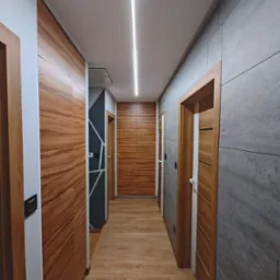 Nowoczesny korytarz z drewnianą zabudową szaf, szarą ścianą z płytek, drewnianymi drzwiami i liniowym oświetleniem LED na suficie.