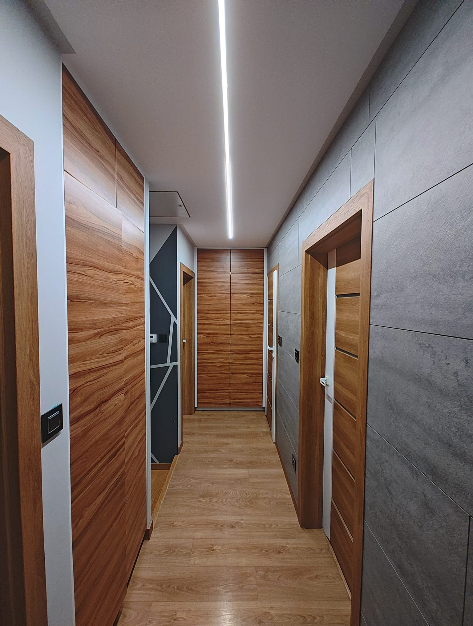 Nowoczesny korytarz z drewnianą zabudową szaf, szarą ścianą z płytek, drewnianymi drzwiami i liniowym oświetleniem LED na suficie.