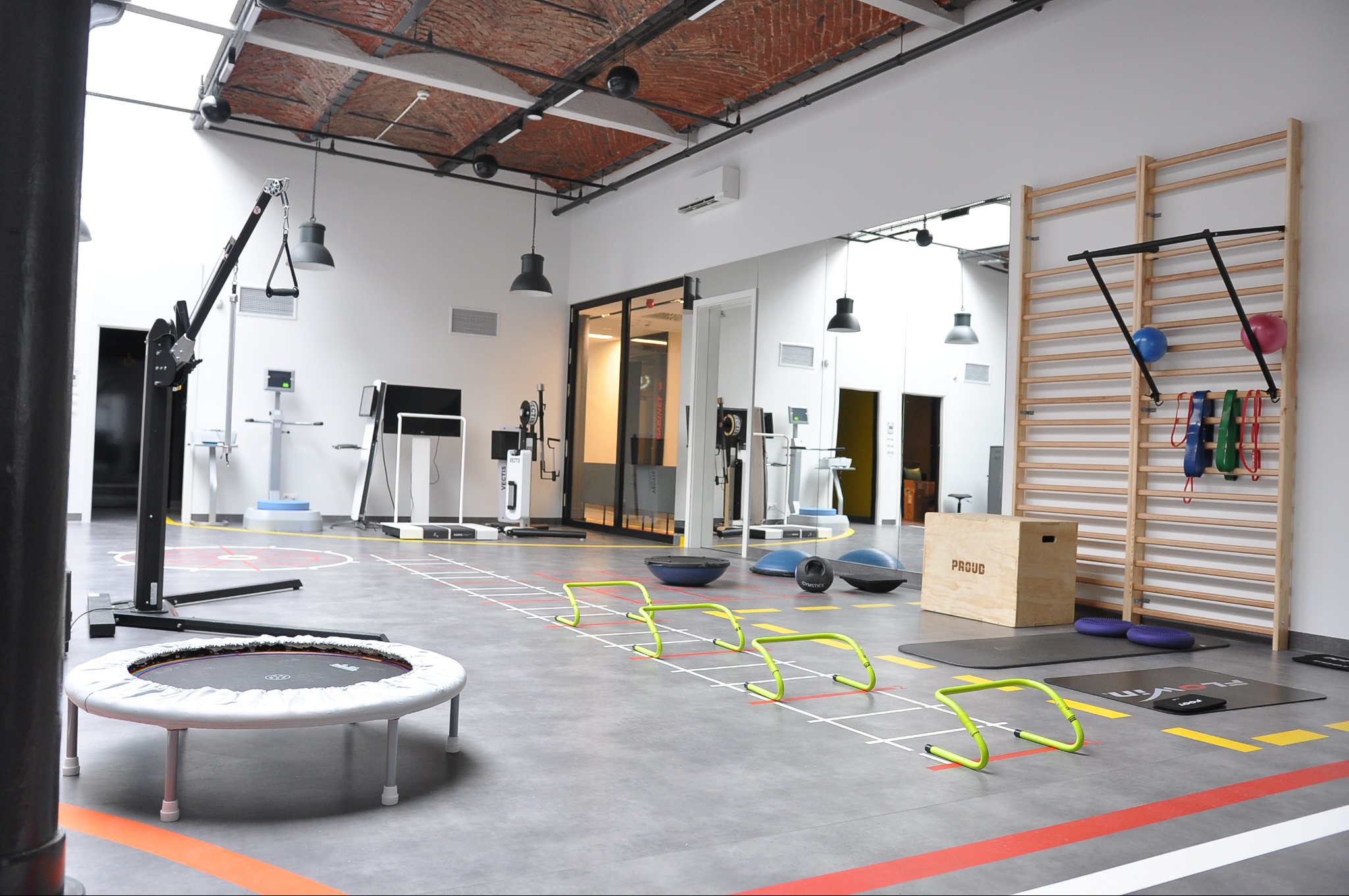 Jasna sala do ćwiczeń z trampoliną, drabinkami gimnastycznymi, niskimi płotkami i innym sprzętem rehabilitacyjnym, widoczny sufit z cegły.