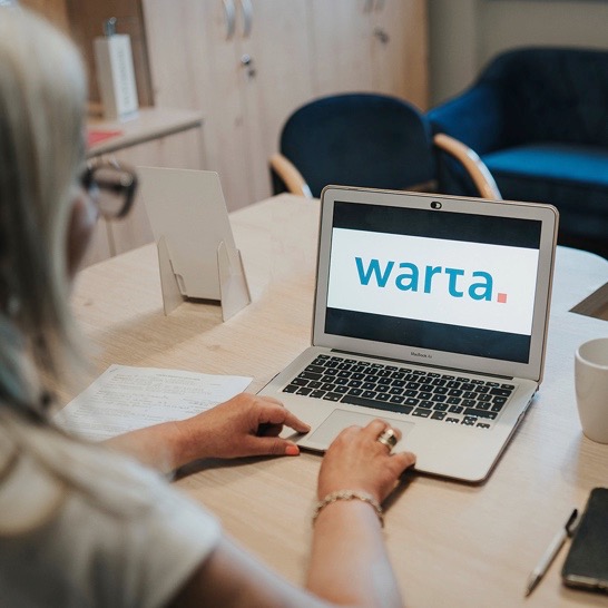 Kobieta przegląda ofertę ubezpieczenia Warta na laptopie w biurze, widoczny dokument, telefon i kubek na biurku.