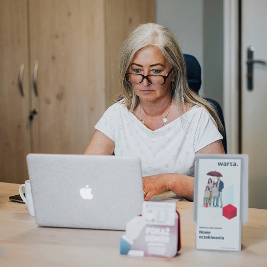 Kobieta w okularach pracuje na laptopie w biurze, obok materiały reklamowe Warta, w tle drewniana szafa.