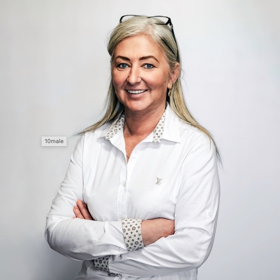 Portret uśmiechniętej kobiety z blond włosami w białej koszuli z wzorzystym kołnierzykiem i mankietami, z okularami na głowie, na jasnym tle.