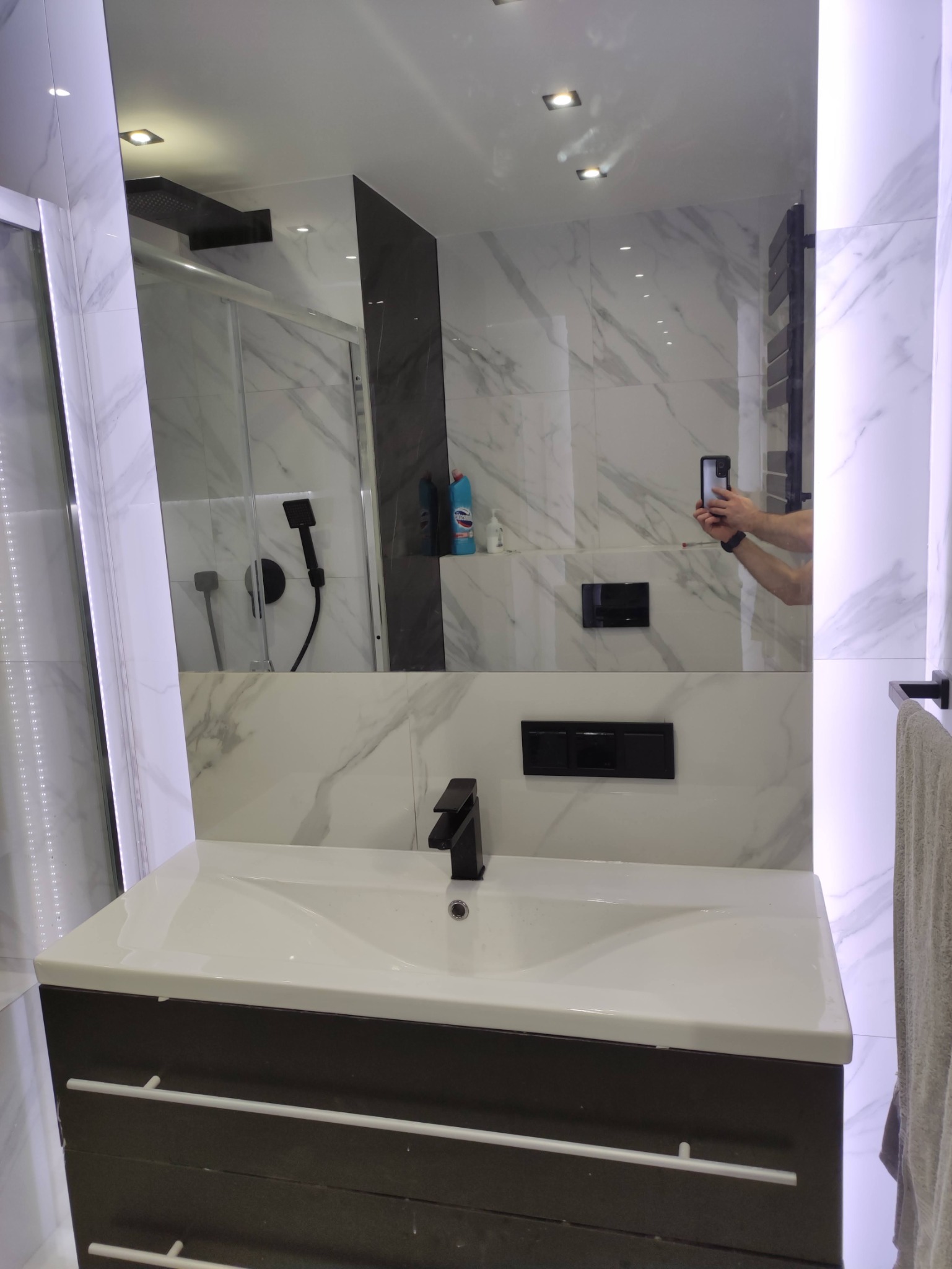 Nowoczesna łazienka w Gdańsku z białymi marmurkowymi płytkami, czarną armaturą i szafką pod umywalką w odcieniach szarości, uchwycona w trakcie wykonywania zdjęcia przez osobę widoczną w lustrze.