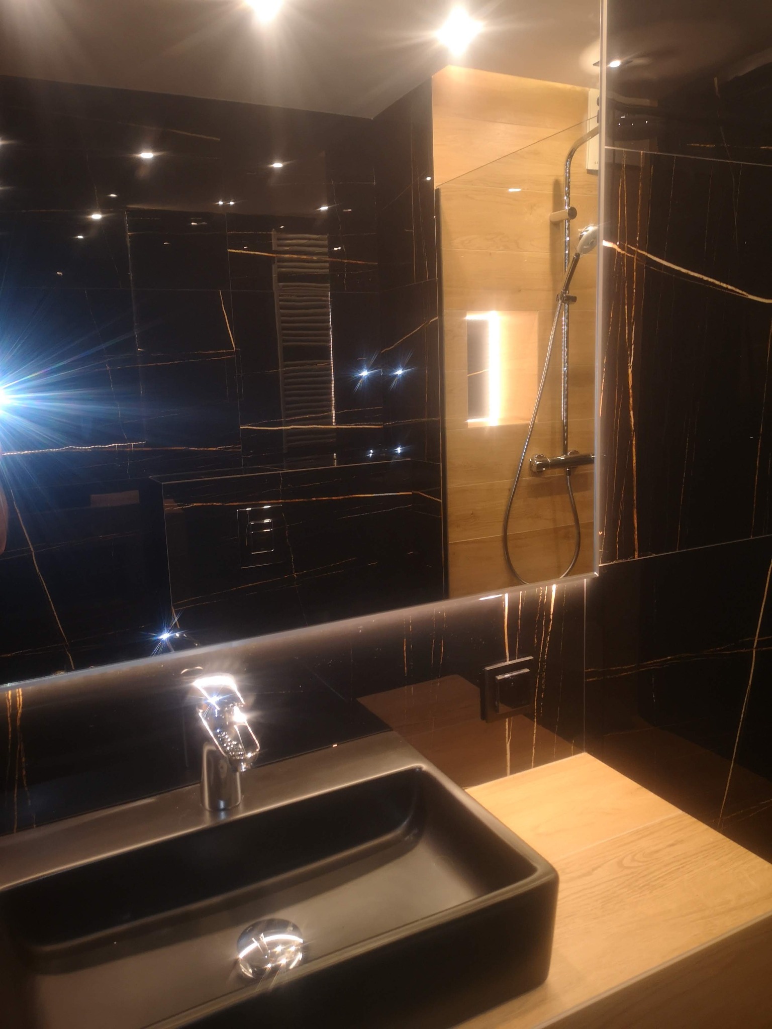 Nowoczesna łazienka z czarnymi płytkami imitującymi marmur z złotymi żyłami, podświetlane lustro z odbiciem kabiny prysznicowej z drewnopodobnymi panelami, czarna umywalka nablatowa z chromowaną...