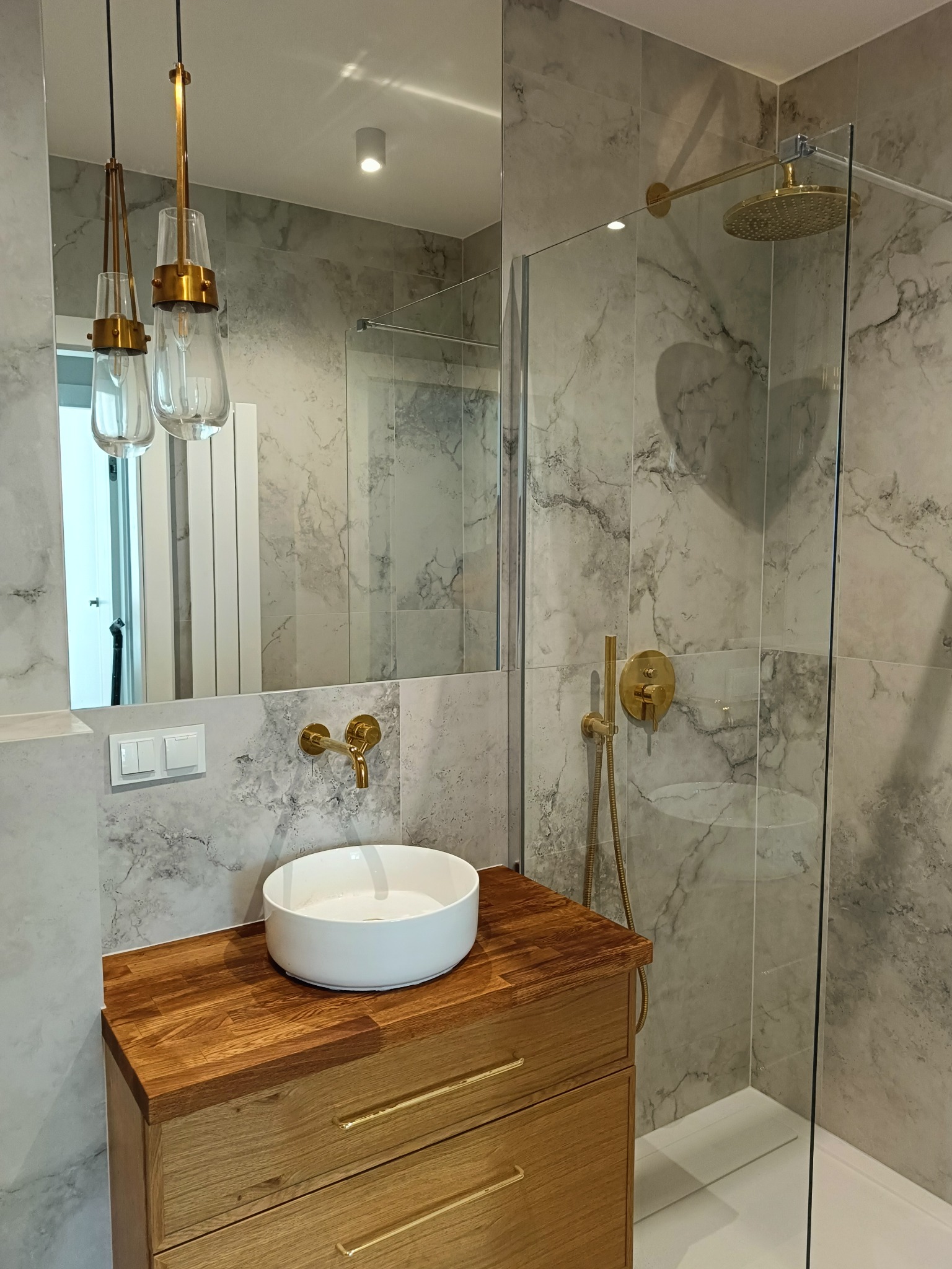 Nowoczesna łazienka z marmurowymi płytkami, złotą armaturą, drewnianą szafką pod umywalką nablatową i szklaną kabiną prysznicową typu walk-in, oświetlona wiszącymi lampami z przezroczystego szkła.