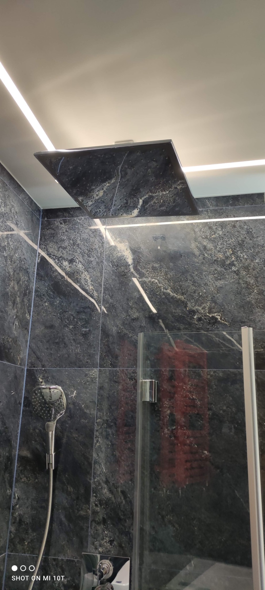 Nowoczesna kabina prysznicowa z deszczownicą i ręczną słuchawką, wykończona czarnym marmurem z białymi żyłami, podświetlana listwą LED w suficie.