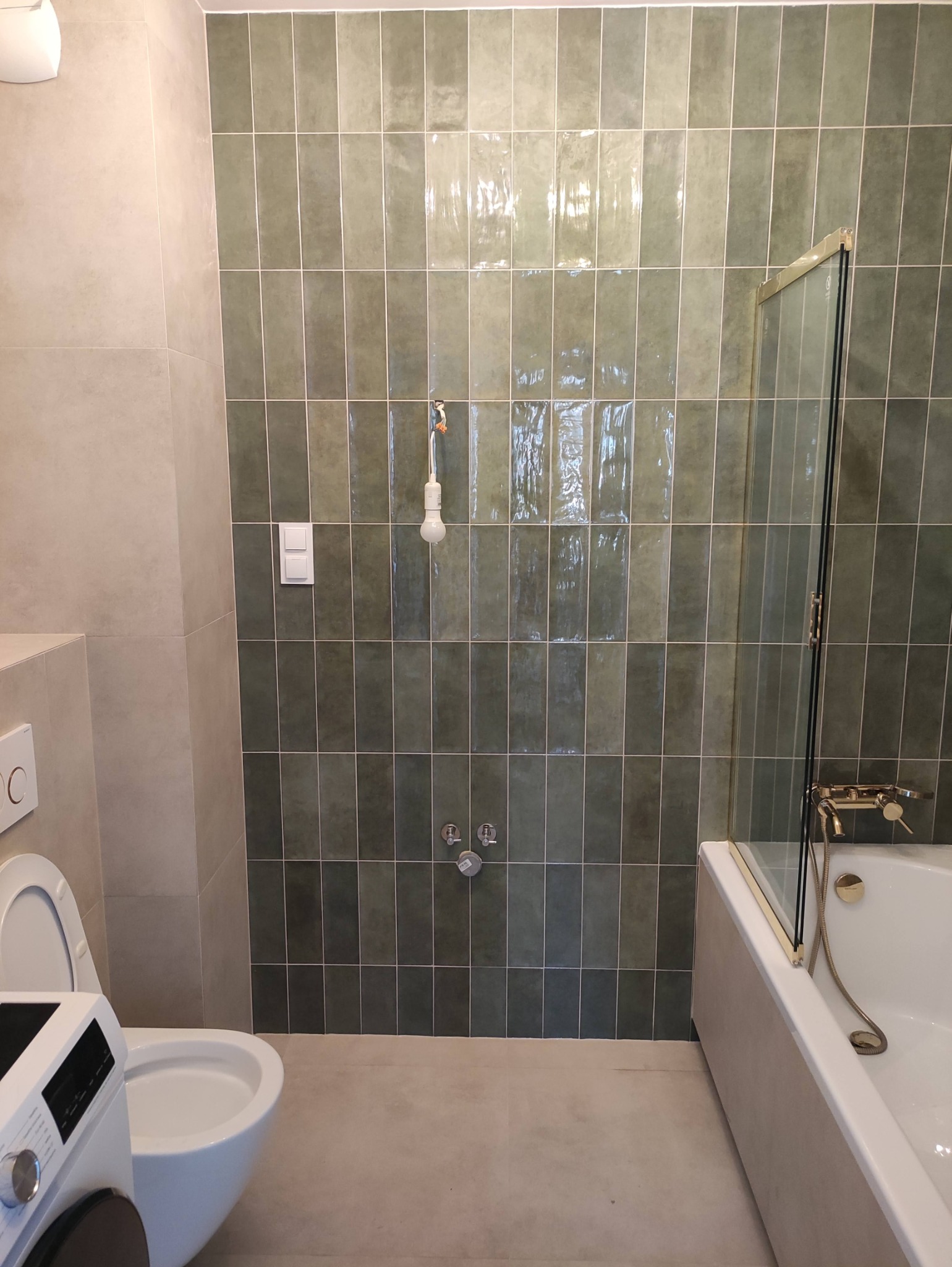 Świeżo wyremontowana łazienka z zielonymi, pionowo ułożonymi płytkami, białą wanną z pozłacaną armaturą i toaletą w rogu, widoczne niedokończone podłączenie oświetlenia.