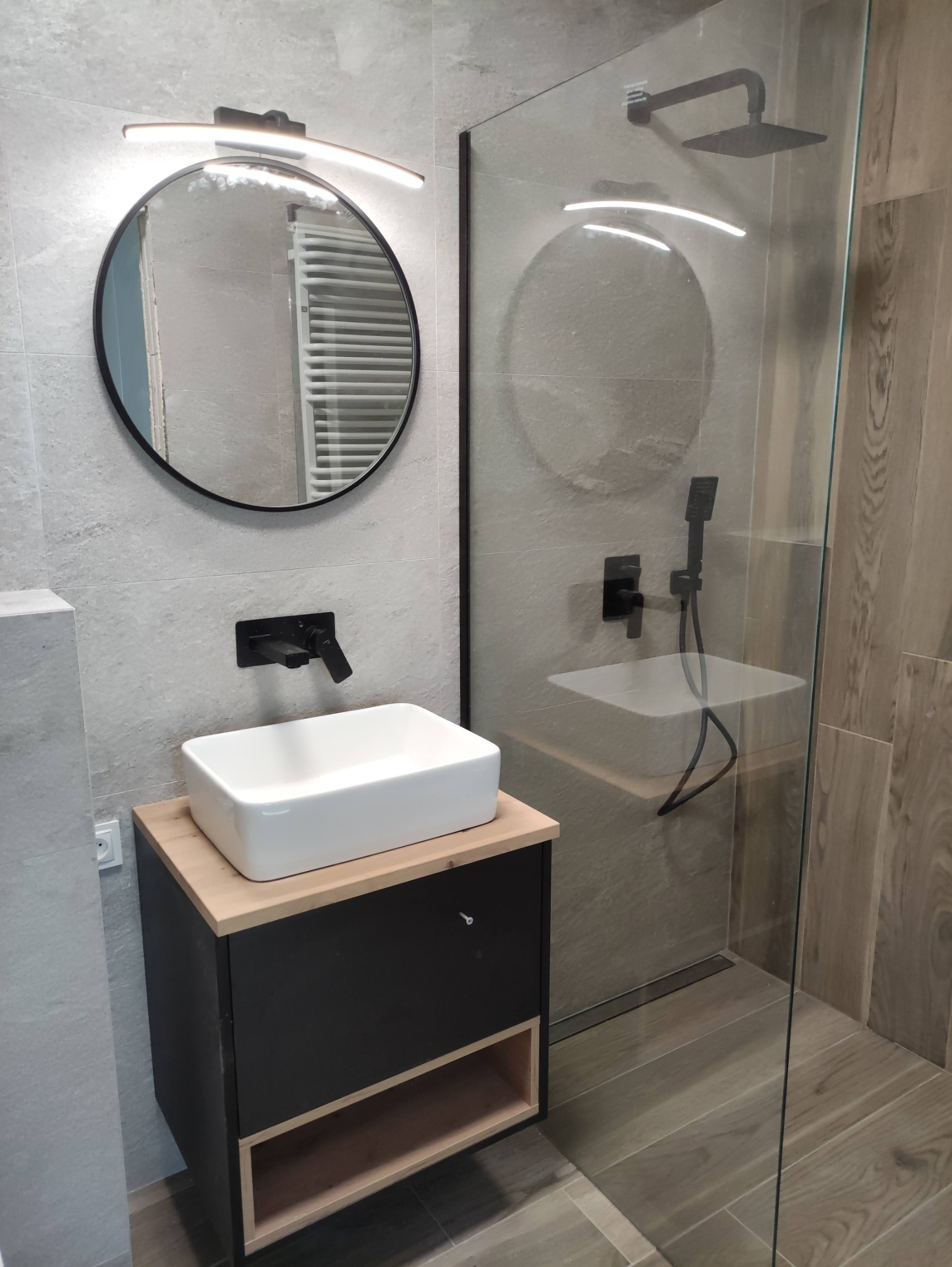 Nowoczesna łazienka z kwadratową umywalką nablatową, czarną armaturą i okrągłym lustrem z podświetleniem LED, kabina prysznicowa typu walk-in z deszczownicą.
