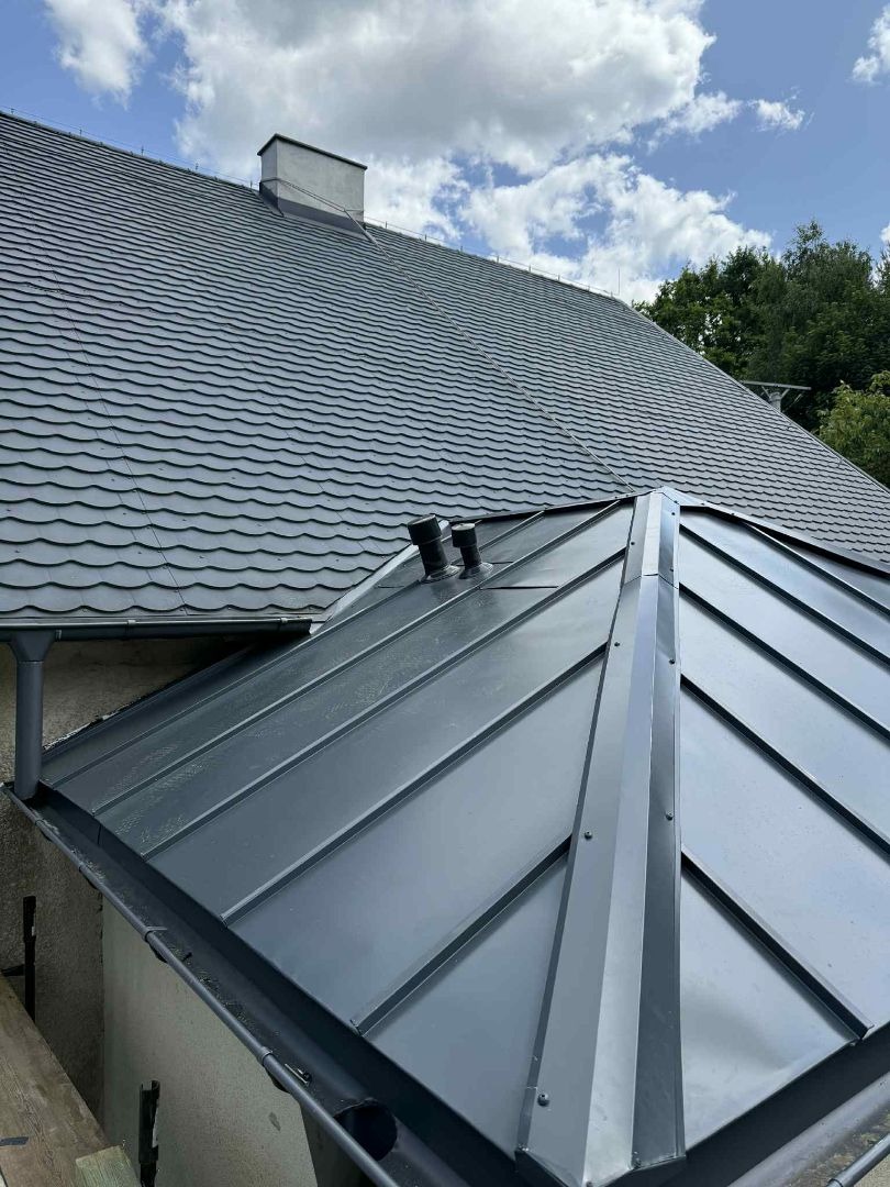 Szary dach z blachy trapezowej z kominem w tle, widok z dołu, rynna i obróbki blacharskie.