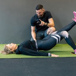 Piotr Piechnik Move Center - Kobieta wykonuje ćwiczenie unoszenia bioder z jedną nogą uniesioną, wspierana przez trenera personalnego w czarnym stroju na zielonej macie w siłowni.