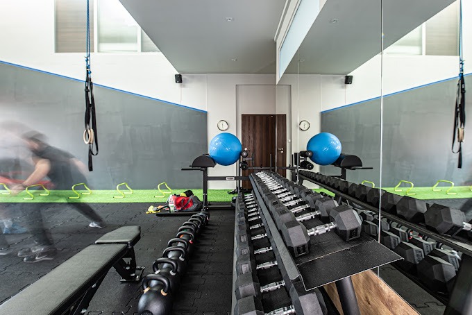 Wnętrze siłowni z rzędem hantli, kettlebells, piłkami gimnastycznymi, drabinkami, ławeczką i rozmazaną sylwetką osoby ćwiczącej w tle.