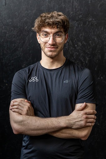 Portret uśmiechniętego mężczyzny w okularach, z założonymi rękami, na ciemnym tle, w sportowej koszulce z widocznymi logotypami.