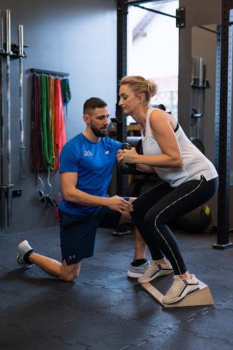 Kobieta wykonuje przysiad z obciążeniem na platformie pod okiem mężczyzny w sportowym stroju w siłowni.