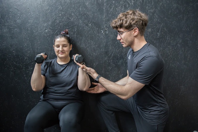 Kobieta siedząca przy ścianie ćwiczy z hantlami, nadzorowana przez mężczyznę w okularach, który koryguje jej postawę.