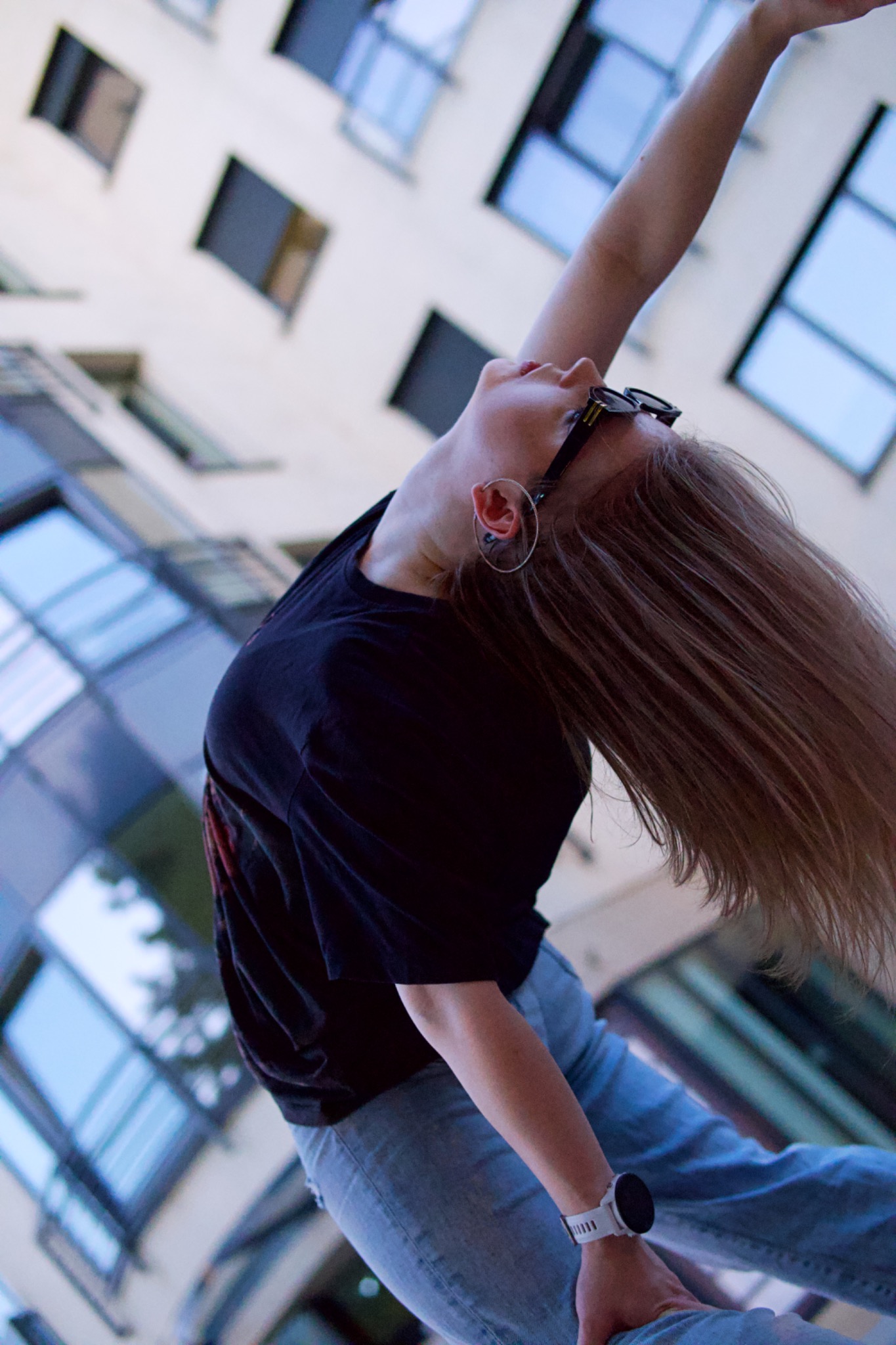 Młoda kobieta w czarnej koszulce i jeansach odchyla głowę do tyłu, z rozwianymi włosami i okularami na głowie, na tle nowoczesnego budynku z licznymi oknami.