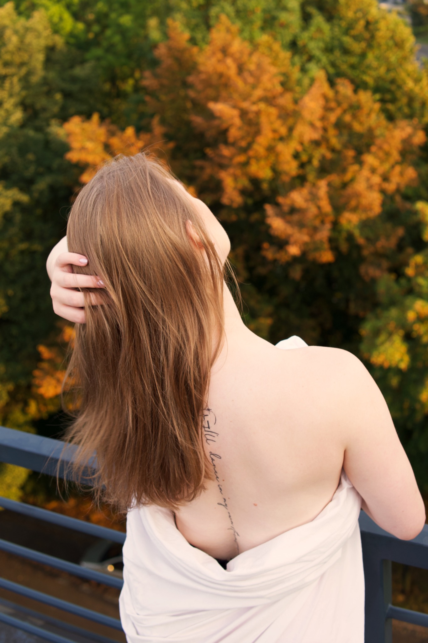 Kobieta z długimi brązowymi włosami, częściowo zakryta białym materiałem, stoi tyłem, patrząc w górę, z widocznym tatuażem na plecach na tle drzew z zielonymi i pomarańczowymi liśćmi.
