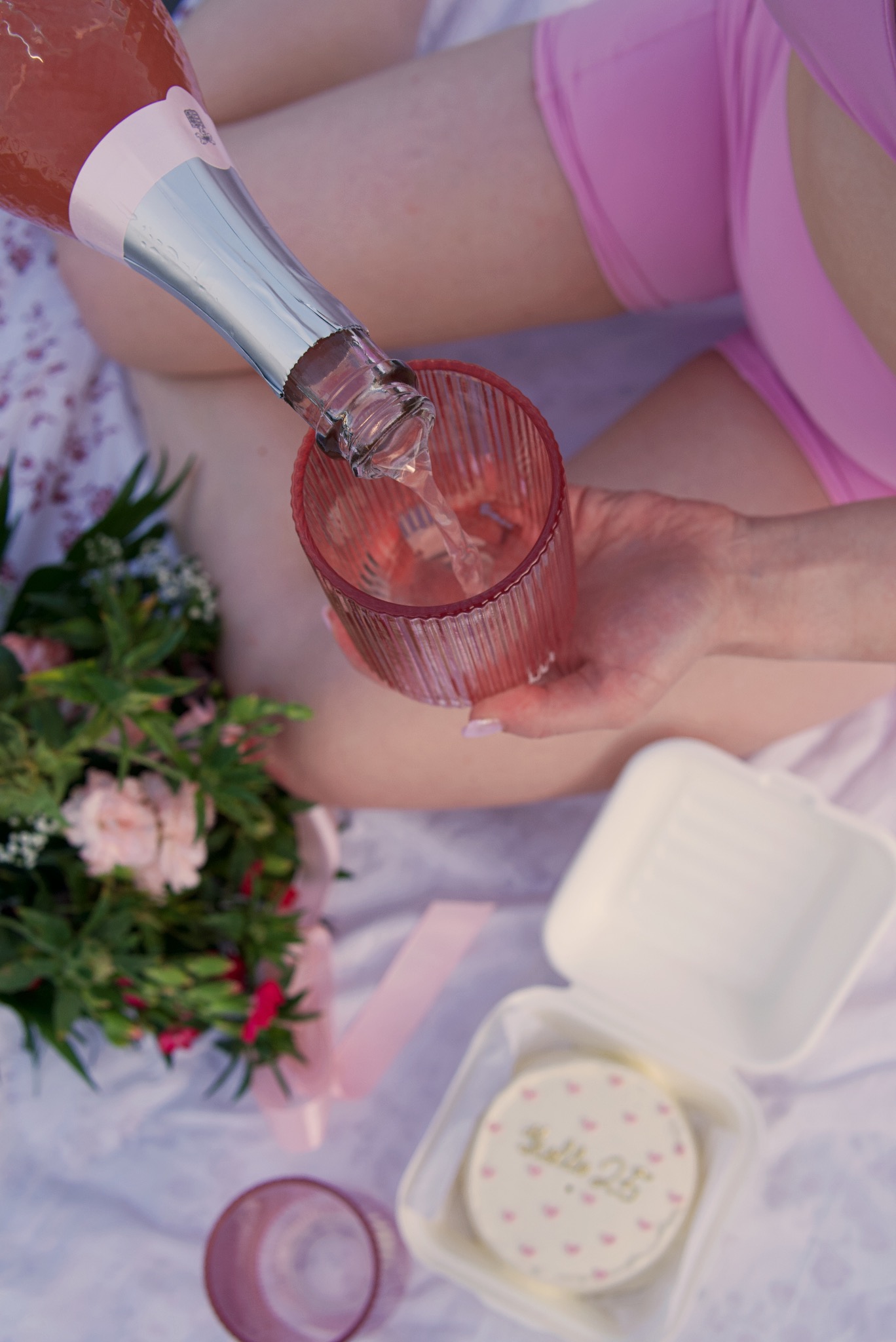 Nalewanie różowego napoju z butelki do szklanki, obok bukiet kwiatów, ciastko w pudełku z napisem 'Hello 25' i różowa wstążka, widok z góry.