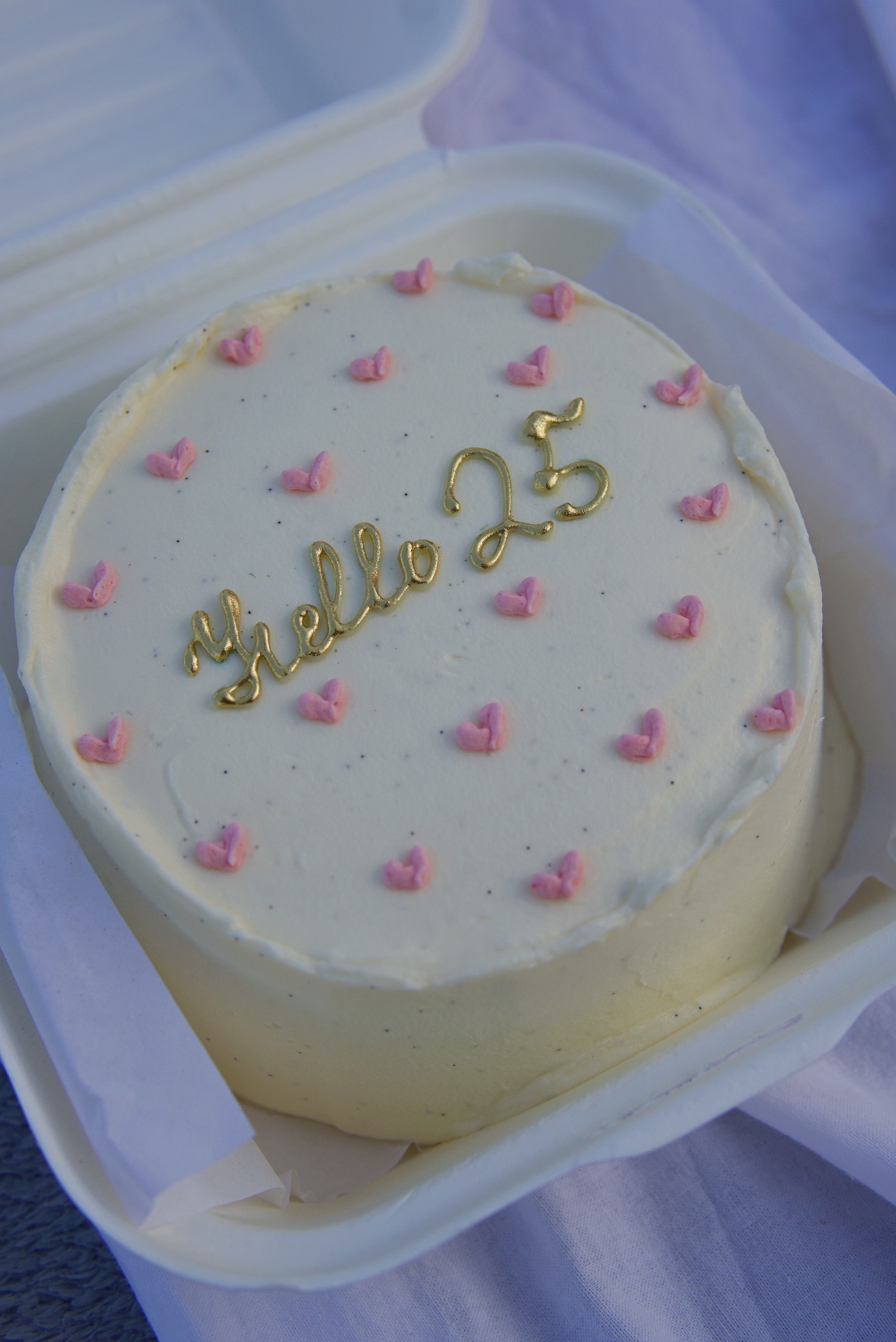 Tort urodzinowy z kremowym lukrem, dekorowany złotym napisem 'Hello 25' i różowymi serduszkami, umieszczony w białym pudełku na tle jasnej tkaniny.