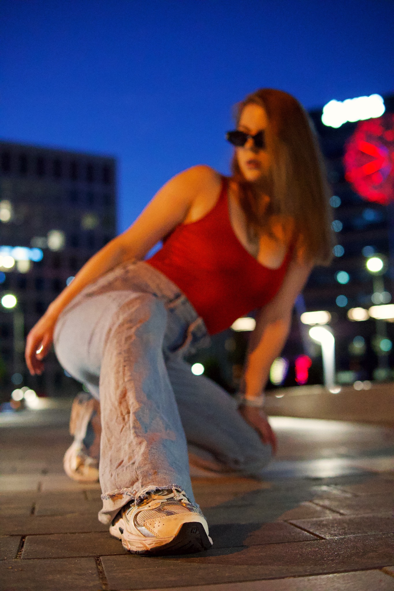 Kobieta w czerwonym topie i jeansach pozuje na tle miejskiego krajobrazu nocą, z ostrością na sportowy but z przodu kadru.