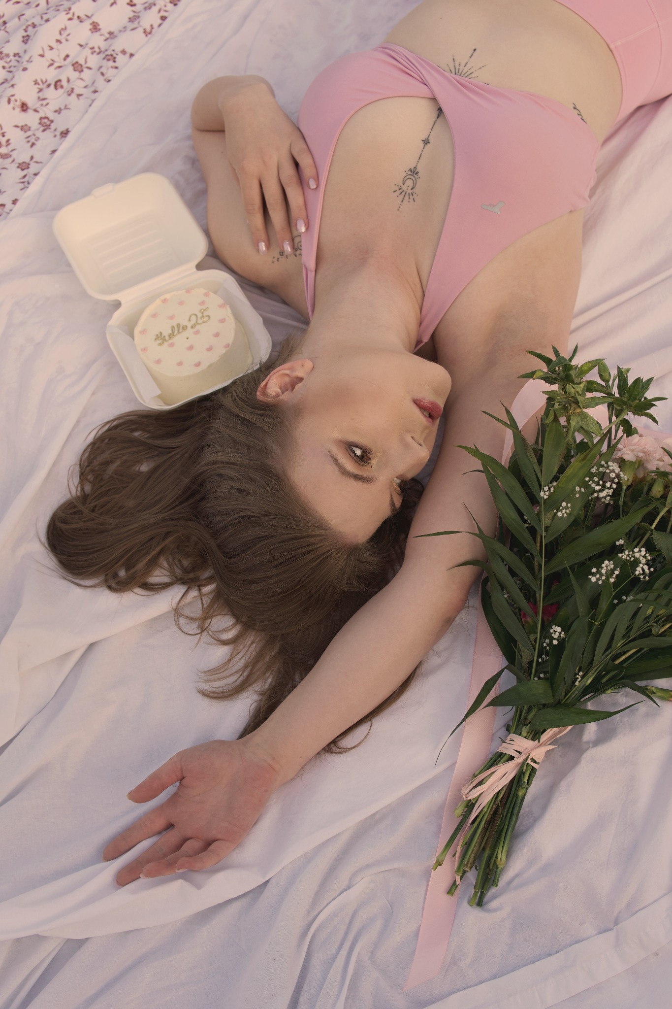 Kobieta leży na białym prześcieradle z bukietem kwiatów i tortem urodzinowym z napisem 'hello 25', ubrana w różowy strój sportowy.