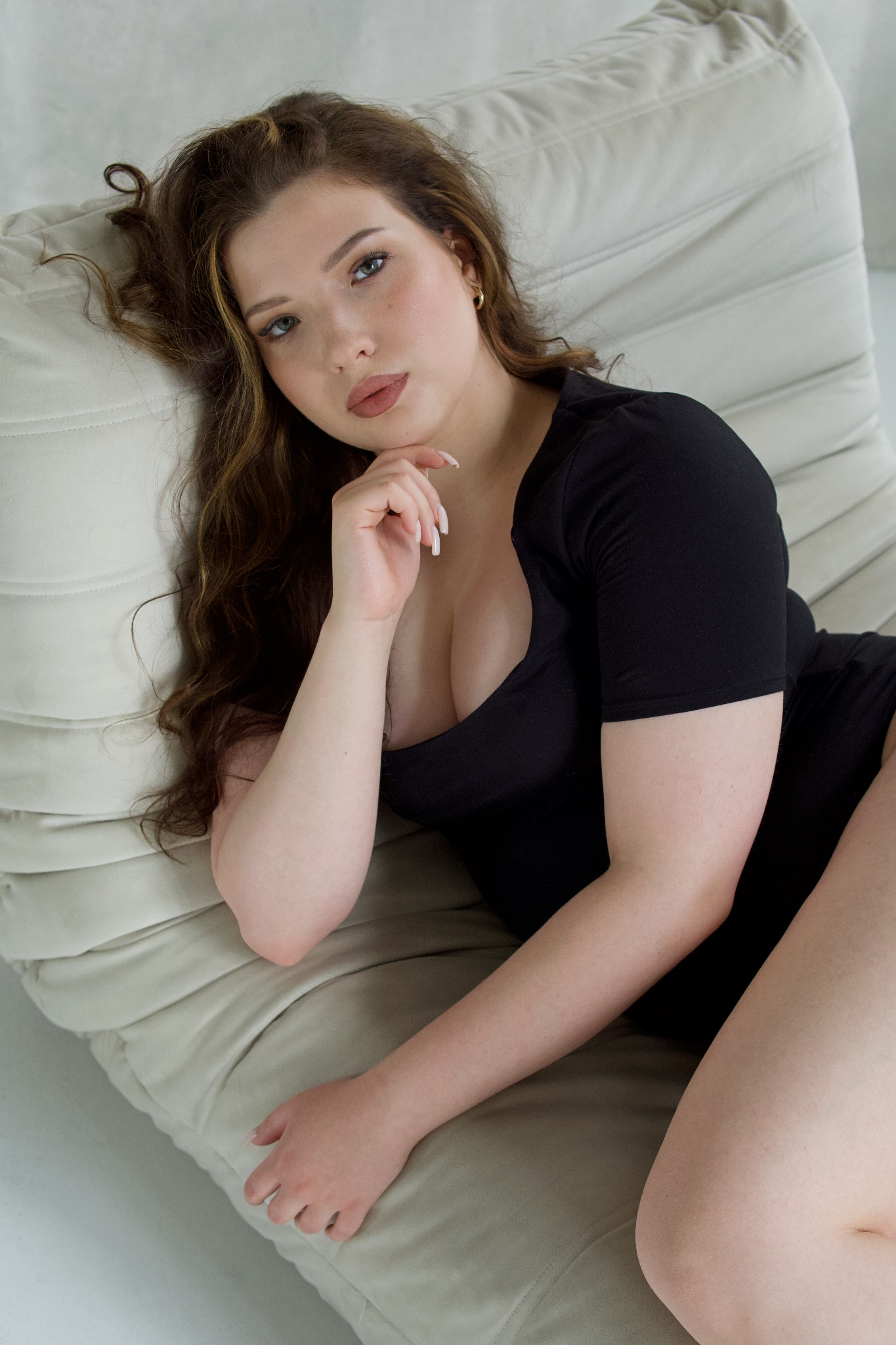Kobieta w czarnej sukience leżąca na beżowej sofie, opierająca podbródek na dłoni z długimi paznokciami, z delikatnym makijażem i złotymi kolczykami.