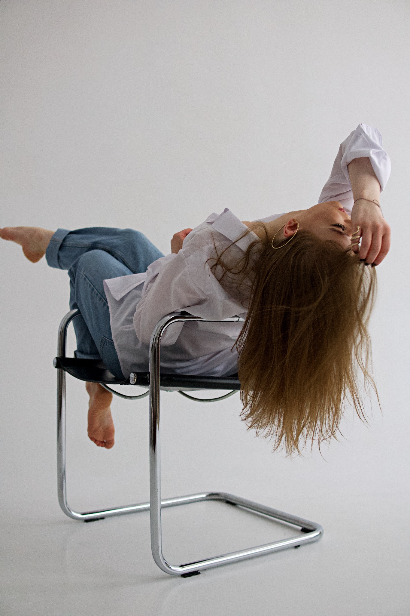 Kobieta w białej koszuli i jeansach leżąca na krześle z chromowanymi nogami na białym tle, z widocznymi złotymi kolczykami kołami.