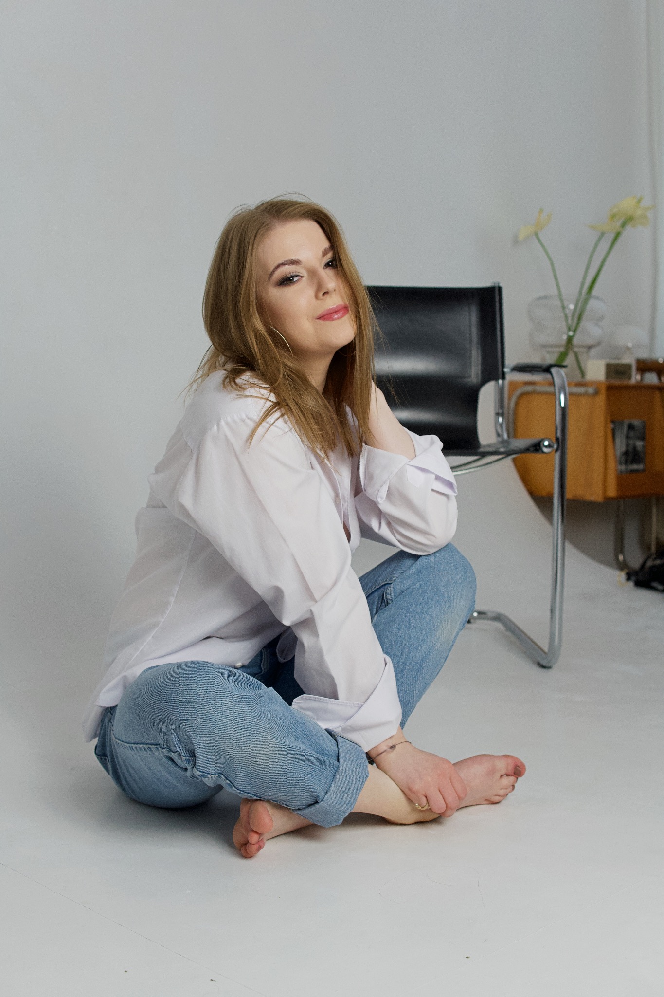 Portret kobiety siedzącej na podłodze w białej koszuli i jeansach, z czarnym krzesłem Thonet i kwiatami w tle
