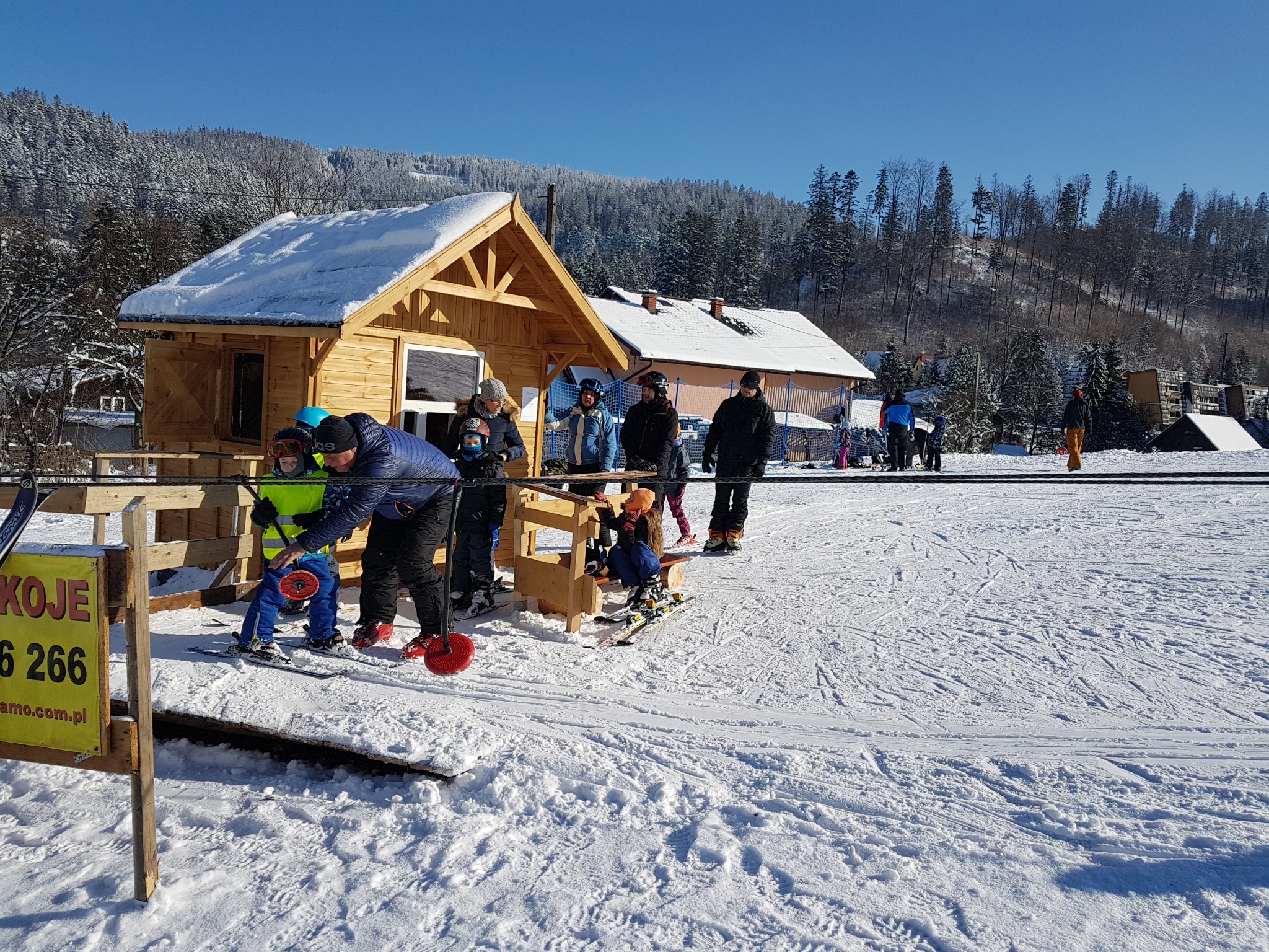 Nauka jazdy na nartach dla dzieci przy wyciągu orczykowym, drewniany budynek w tle, zimowy krajobraz z zalesionymi wzgórzami.