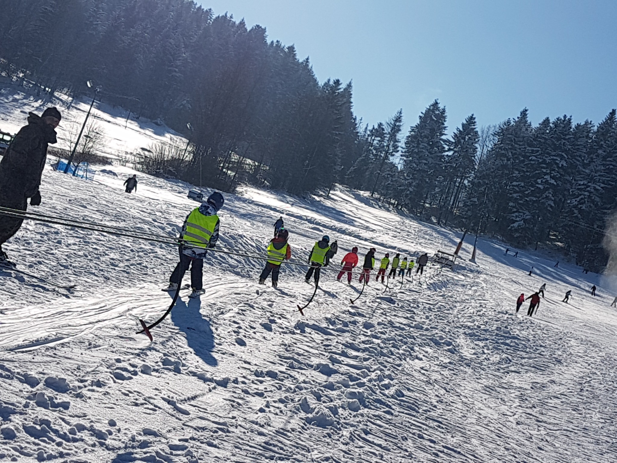 Grupa dzieci z instruktorami w jaskrawych kamizelkach wjeżdża wyciągiem orczykowym na ośnieżony stok narciarski w słoneczny dzień, w tle las iglasty.