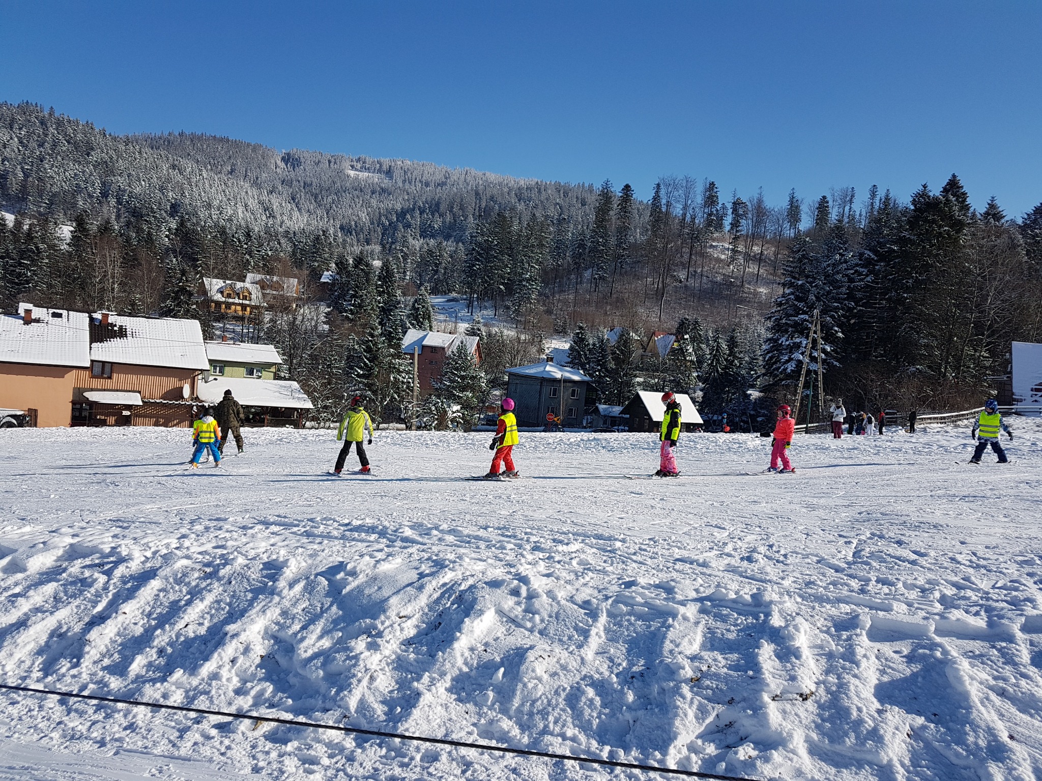 Grupa dzieci uczy się jeździć na nartach na ośnieżonym stoku w słoneczny dzień, w tle widoczne budynki i zalesione wzgórze.