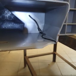 SKEMWELD PRZEDSIĘBIORSTWO WIELOBRANŻOWE PIOTR KUSZYŃSKI - Szara plastikowa skrzynka z pęknięciem naprawionym czarnym klejem, widoczna na tle metalowego stelaża i kafelkowej podłogi.