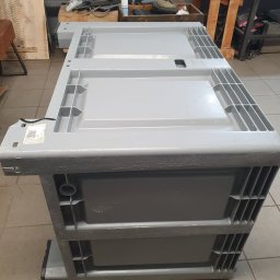 SKEMWELD PRZEDSIĘBIORSTWO WIELOBRANŻOWE PIOTR KUSZYŃSKI - Szara, plastikowa skrzynia transportowa z ażurowymi ściankami, widziana z perspektywy żabiej, na tle warsztatu.