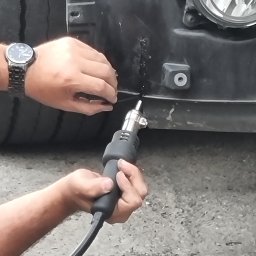 SKEMWELD PRZEDSIĘBIORSTWO WIELOBRANŻOWE PIOTR KUSZYŃSKI - Naprawa plastikowego zderzaka samochodowego za pomocą opalarki; widoczne ręce, zegarek na nadgarstku, fragment opony i reflektora.