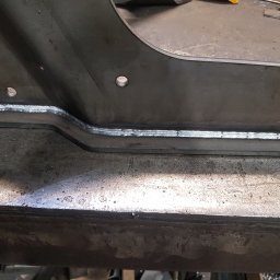 SKEMWELD PRZEDSIĘBIORSTWO WIELOBRANŻOWE PIOTR KUSZYŃSKI - Metalowy element konstrukcyjny po spawaniu, widoczne spawy pachwinowe z charakterystycznym wzorem, trzy otwory montażowe, całość na warsztatowym stole.