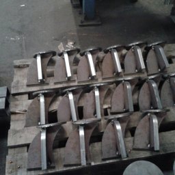 SKEMWELD PRZEDSIĘBIORSTWO WIELOBRANŻOWE PIOTR KUSZYŃSKI - Rząd elementów metalowych, prawdopodobnie wsporników, ułożonych na drewnianej palecie w warsztacie, z widocznymi śladami obróbki.