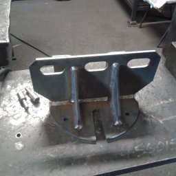 SKEMWELD PRZEDSIĘBIORSTWO WIELOBRANŻOWE PIOTR KUSZYŃSKI - Metalowy wspornik spawany z blachy i prętów, widoczne spawy, leżący na metalowym stole warsztatowym obok narzędzi