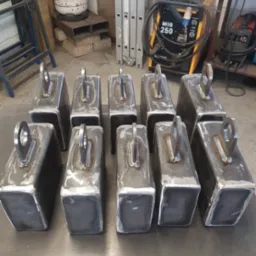 Partia spawanych, surowych, stalowych elementów w kształcie prostopadłościanów z przyspawanymi uchwytami na płaskiej powierzchni roboczej, w tle spawarka MIG 250 i drabina.