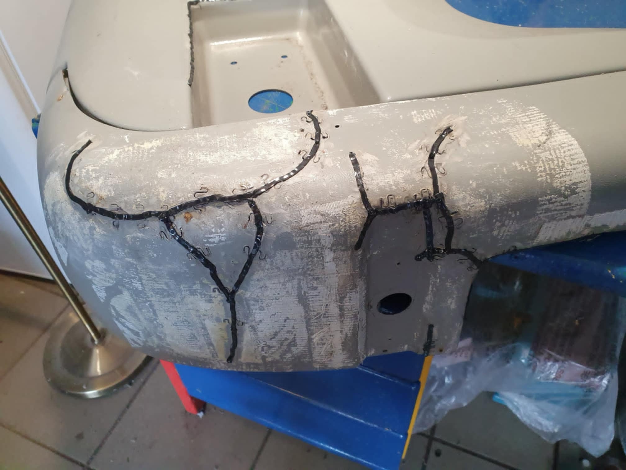 Szary element konstrukcyjny z metalu, naprawiany za pomocą zszywek i czarnego kleju, widoczne ślady szlifowania i przygotowania do malowania.